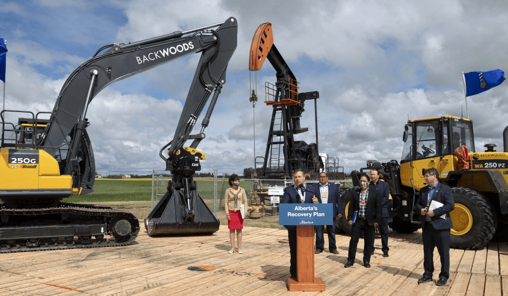 A picture of former Alberta Premier, Jason Kenney, announcing the SRP funding program. The picture includes some environmental reclamation equipment in the background, and Mr. Kenney along with 5 other people are standing on a stage. Mr. Kenney is speaking at a podium. The podium has a sign on the front that says ' Alberta's Recovery Plan' and has an Alberta Government logo.